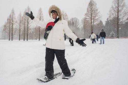 A person in a white coat and fur hood joyfully snowshoes in a snowy park, with others walking in the background.