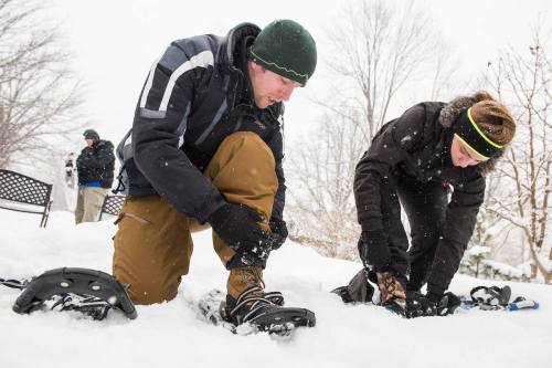 Two people in winter gear adjusting snowshoes in a snowy landscape, with trees and a person in the background.