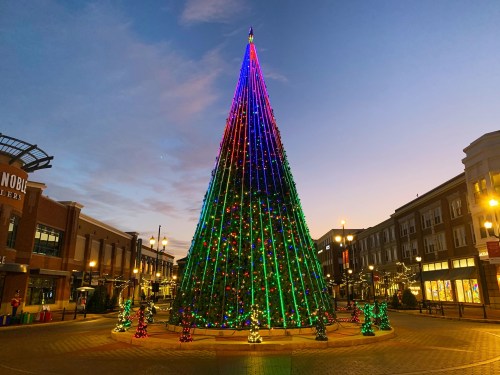 A brightly lit Christmas tree adorned with colorful lights stands in a plaza at dusk, surrounded by festive decorations.