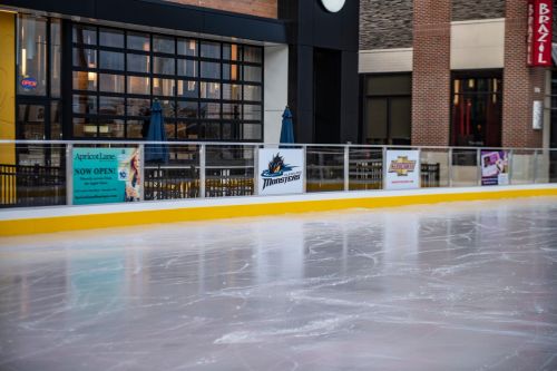 An empty ice rink with a yellow border, featuring signs for local businesses and a view of nearby seating areas.