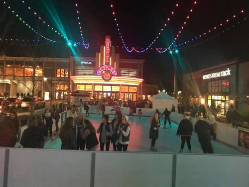 People ice skating at night in front of a cinema, with colorful lights and shops in the background.