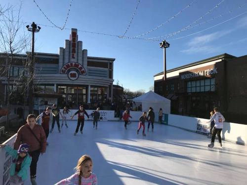 Children and adults ice skating on a rink outside a cinema, with festive lights and a clear blue sky in the background.