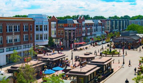 A vibrant town square with shops, outdoor seating, and people enjoying a sunny day under a blue sky.