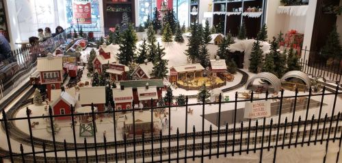 A festive model train display featuring a snowy village, trees, and a train track surrounded by a decorative fence.