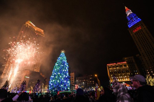 A festive scene with a large illuminated Christmas tree, fireworks, and a city skyline adorned with holiday lights.