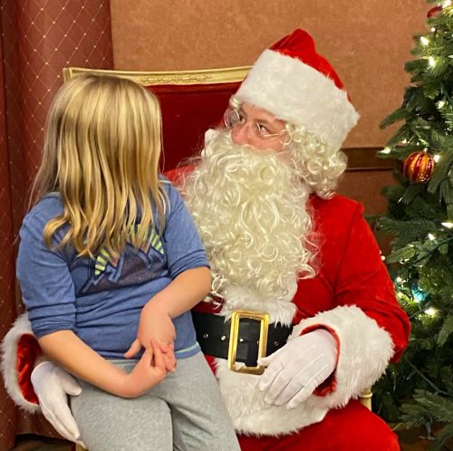 A child sits on Santa's lap, smiling and looking up at him, with a decorated Christmas tree in the background.