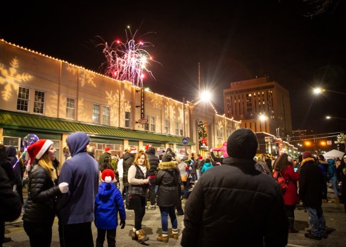 Crowd gathered at night for a festive event, with fireworks lighting up the sky above decorated buildings.