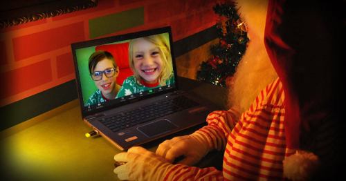 Santa in a red suit video chatting with two smiling children on a laptop, festive decorations in the background.