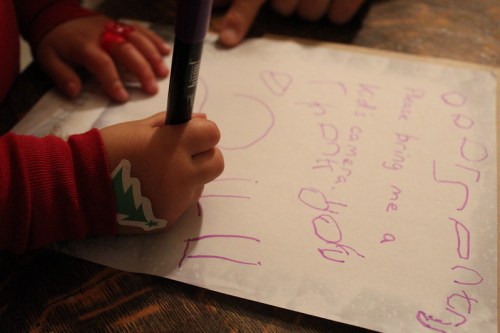 A child's hand holding a marker, writing a note on paper with colorful letters and a festive sticker on their wrist.