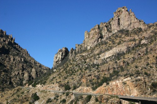 A winding road through rugged mountains under a clear blue sky, showcasing steep rock formations and dry vegetation.