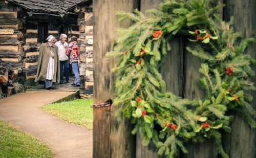 A festive wreath hangs on a door as two people chat with a person in historical attire near a log cabin.