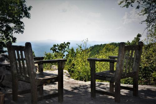 Two rustic wooden chairs overlook a scenic mountain view, surrounded by greenery under a bright sky.