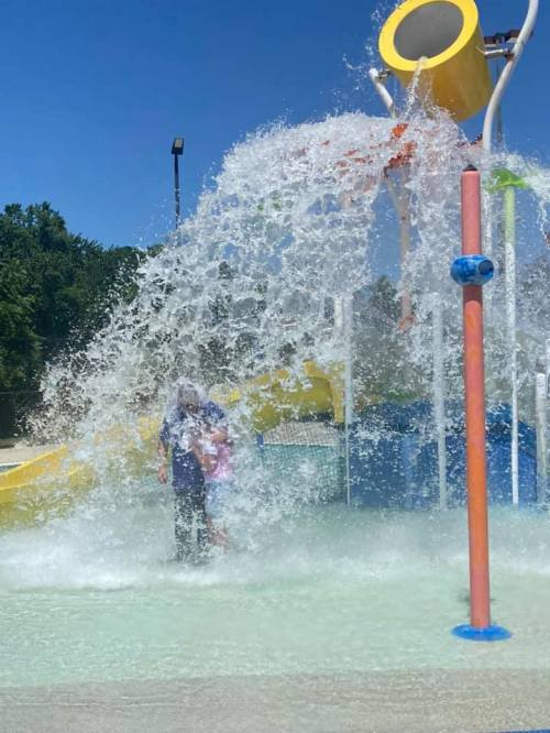 A child and adult stand under a large splash of water at a water park, with slides and colorful structures in the background.