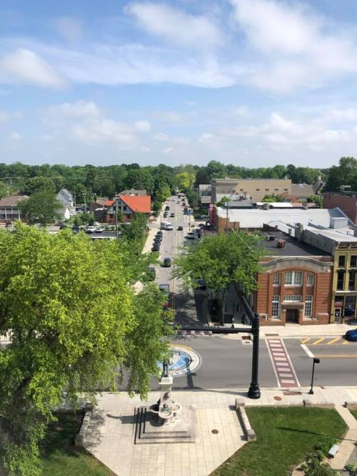 Aerial view of a tree-lined street with shops and cars, under a blue sky with scattered clouds.