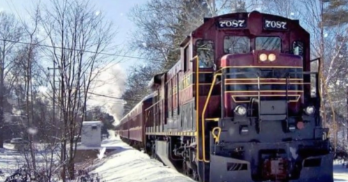 A vintage train travels through a snowy landscape, surrounded by trees and a small building in the background.