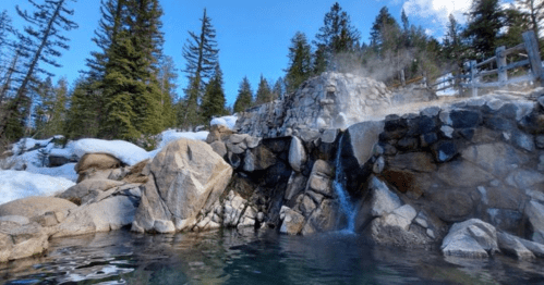 A serene hot spring surrounded by snow-covered rocks and tall pine trees under a clear blue sky.