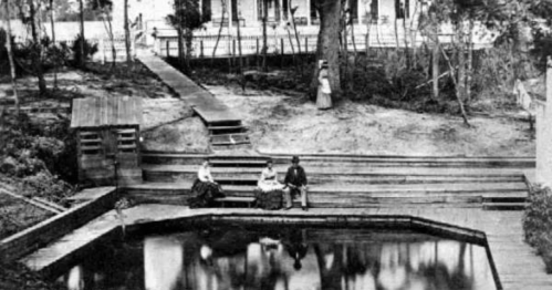 Black and white photo of a garden with a pond, featuring three people seated and a woman standing in the background.