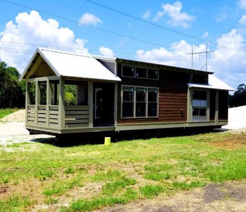 A modern tiny house with a porch, featuring a mix of wood and metal siding, set against a blue sky with clouds.