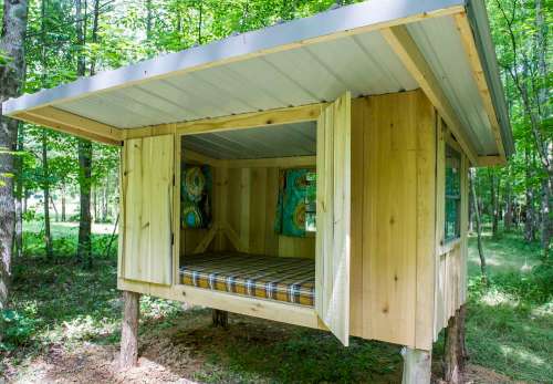 A small wooden cabin on stilts in a forest, featuring open windows and a bed with a checkered blanket inside.