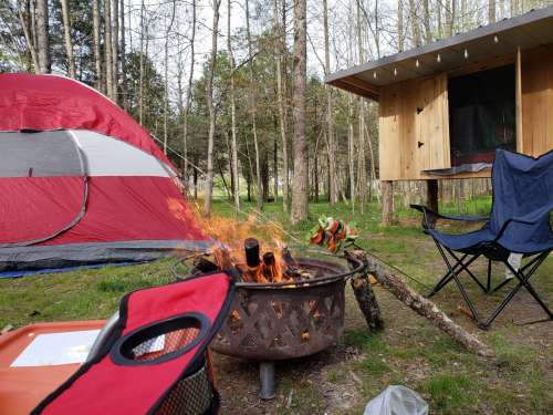 A campsite with a red tent, a fire pit with flames, and camping chairs surrounded by trees.