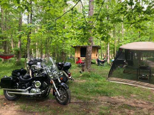 A motorcycle parked near a campsite with a tent, hammock, and people relaxing among green trees.