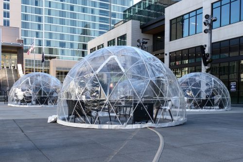 Three transparent geodesic domes set up on a paved area, surrounded by modern buildings and outdoor lighting.