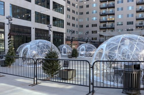 Clear geodesic domes set up in a courtyard, surrounded by fences and small trees, with buildings in the background.