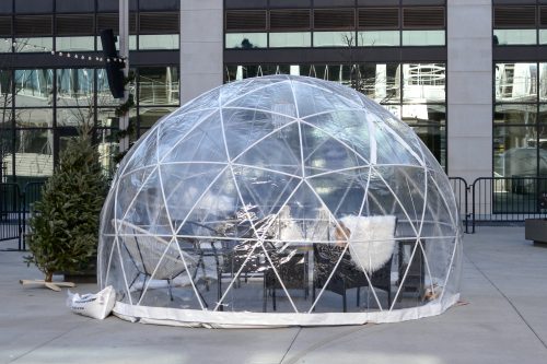 A transparent geodesic dome with seating inside, set in an urban area with buildings in the background.
