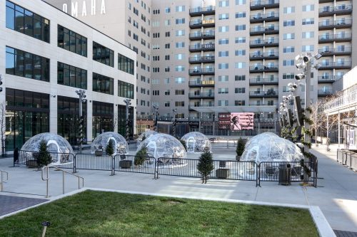 A courtyard with clear dome structures, surrounded by greenery and modern buildings, featuring a large screen in the background.