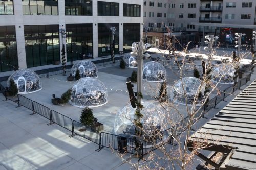 Aerial view of transparent igloo-like structures in an urban plaza, surrounded by trees and barriers.