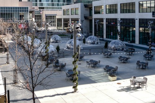 Outdoor plaza with geodesic domes, tables, and string lights, surrounded by modern buildings and bare trees.
