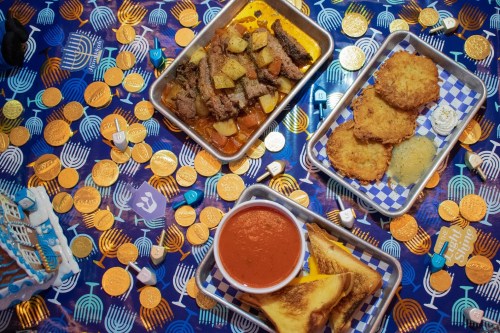 A festive table with trays of brisket, latkes, tomato soup, and gelt coins, celebrating a holiday.