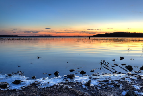 A serene lake at sunset, with calm waters reflecting vibrant orange and blue hues, surrounded by rocks and sparse vegetation.