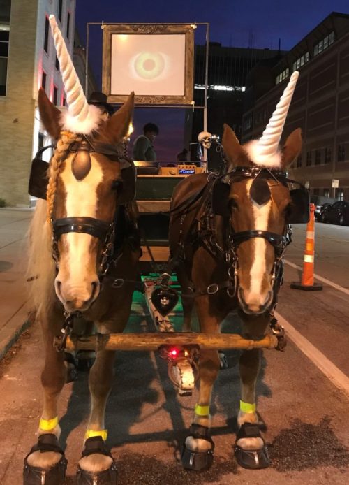 Two horses with unicorn horns stand side by side, harnessed to a carriage, against a twilight city backdrop.