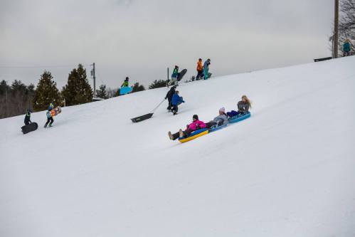 Children and adults sledding down a snowy hill, with some walking up carrying sleds in the background.