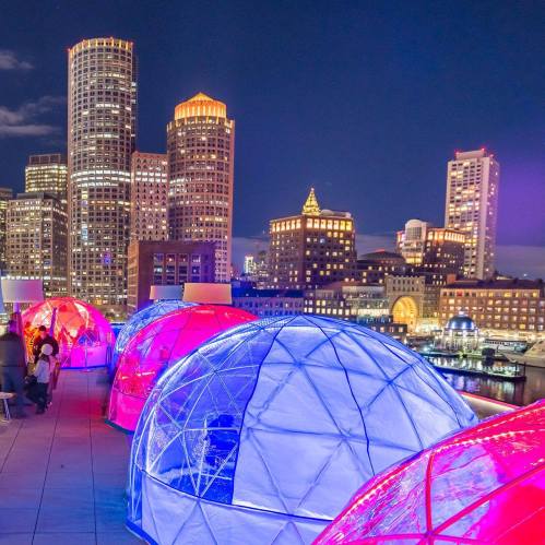 Colorful illuminated igloos on a rooftop with a city skyline at night in the background.