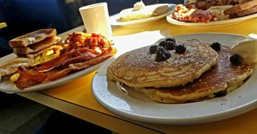 A plate of pancakes topped with blueberries, alongside bacon, toast, and a cup of coffee on a yellow table.
