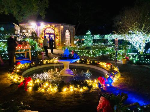 A festive outdoor scene at night, featuring a fountain surrounded by colorful lights and holiday decorations.