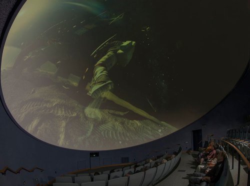A planetarium interior with an audience watching a space-themed projection on a domed ceiling.