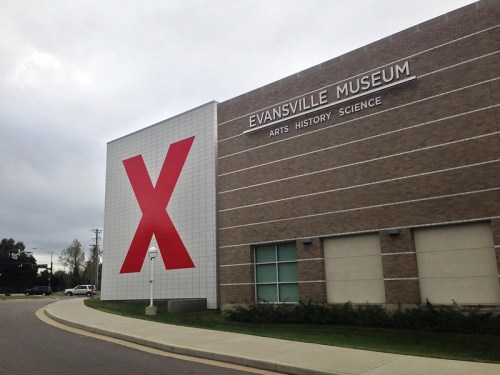 Exterior of the Evansville Museum featuring a large red "X" on the wall, with a cloudy sky in the background.