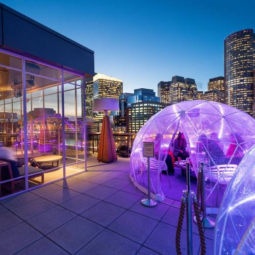 A rooftop terrace with glowing igloos, city skyline at dusk, and modern seating under a twilight sky.