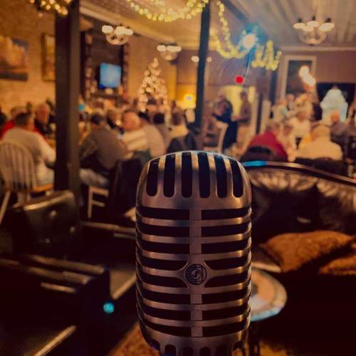 A vintage microphone in the foreground with a lively crowd and festive decorations in a cozy venue.