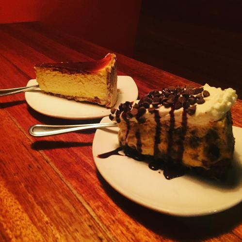 Two slices of cheesecake on white plates, one topped with caramel and the other with chocolate chips and whipped cream.