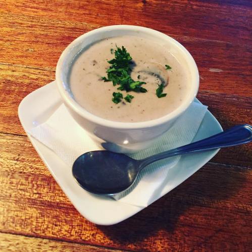 A bowl of creamy soup garnished with herbs, served on a white plate with a spoon beside it on a wooden table.