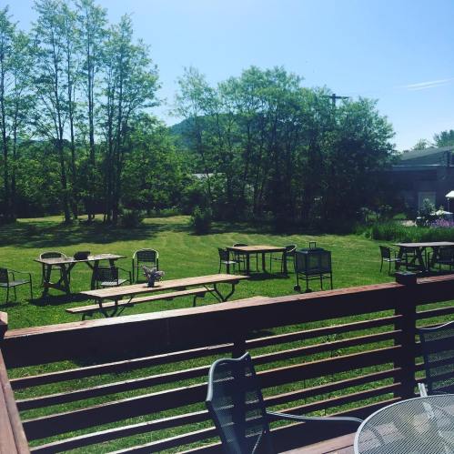 A sunny outdoor area with empty tables on a grassy lawn, surrounded by trees and distant hills.