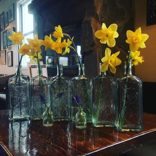 A collection of vintage glass bottles with yellow daffodils and purple flowers arranged on a wooden table.