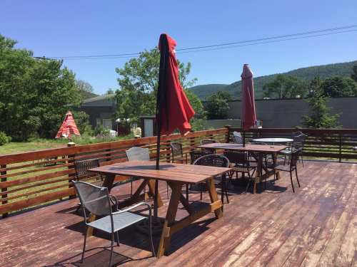 A sunny outdoor deck with wooden tables, chairs, and red umbrellas, surrounded by greenery and distant hills.