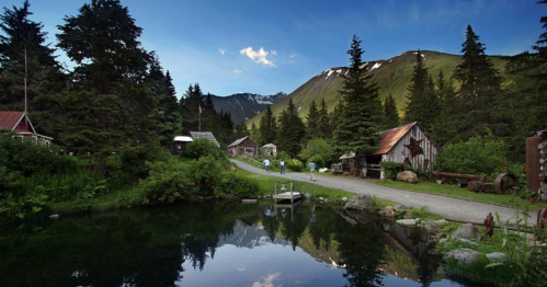 A serene landscape featuring a dirt road, rustic buildings, tall trees, and a calm pond with mountains in the background.