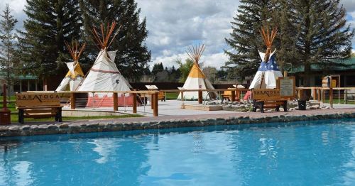 A serene outdoor scene featuring tipis near a clear blue pool, surrounded by trees and a cloudy sky.