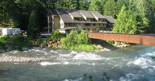 A riverside lodge surrounded by trees, with a bridge crossing over a flowing river and a camper nearby.